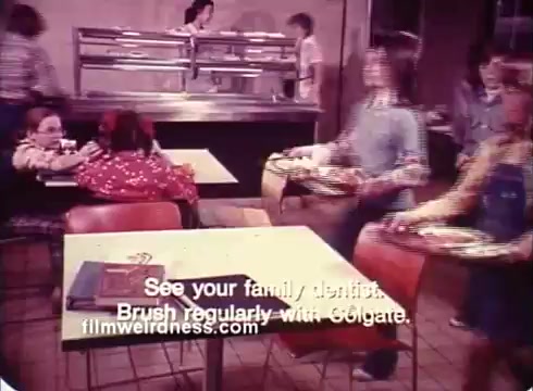 The still depicts a scene from a Colgate TV commercial featuring children, some wearing braces, in a cafeteria setting. A few kids are seated at tables, while others are walking by with food trays. The text at the bottom encourages viewers to visit their dentist and brush regularly with Colgate, emphasizing dental hygiene. The overall atmosphere is lively and focused on promoting good oral care habits among children.