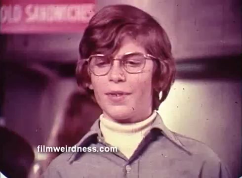 The still from the Colgate TV commercial features a young boy with glasses and braces, wearing a turtleneck under a collared shirt. He appears to be speaking, likely in a friendly or engaging manner. The background includes a sign with the words 'COLD SANDWICHES,' suggesting a casual setting, possibly a diner or snack area. The overall tone is nostalgic, typical of advertisements aimed at children and promoting dental care.