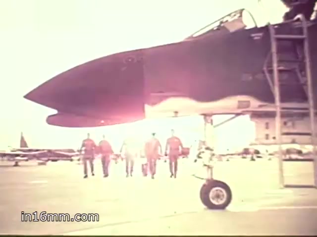 The still captures a moment from a 1968 Vietnam-era Air Force ROTC recruiting commercial featuring an F-4 Phantom II. In the foreground, the aircraft's nose is prominent, basking in bright sunlight that creates a glowing effect. A group of military personnel walks confidently towards the camera, suggesting camaraderie and a sense of duty. Their uniforms indicate active service roles, while the aircraft in the background highlights the technological prowess of the Air Force. The scene conveys themes of recruitment, patriotism, and the allure of military service during a turbulent period in U.S. history.