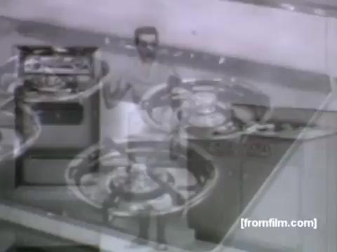 The still features a vintage kitchen setting from the 1950s, prominently displaying a Caloric gas range and oven. In the foreground, a man, likely the host of the commercial, is demonstrating the appliance, possibly showcasing its features or functionality. The overall style reflects the clean lines and aesthetic typical of UPA animation, focusing on modern convenience in home cooking. The scene conveys a sense of innovation and domestic ease, characteristic of advertisements from that era.