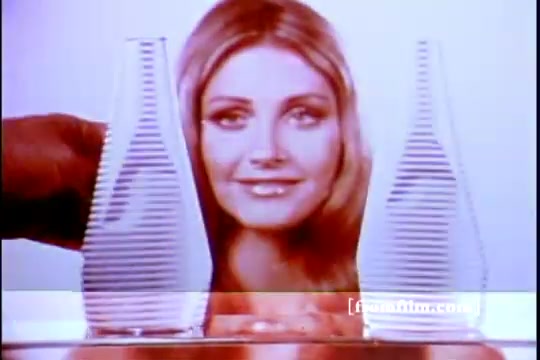 The still from the 1970s 'Twice as Nice' shampoo commercial features a woman with long, blonde hair and a bright smile, positioned between two clear, striped bottles. The background is simply designed to keep the focus on the woman and the product. The overall aesthetic reflects the vibrant and stylized visuals typical of that era in advertising.