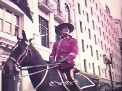 The still features a mounted Royal Canadian Mounted Police officer in traditional red serge, riding through an urban setting, likely a downtown area. The officer's attire and the horse add a nostalgic touch, emphasizing national identity and heritage. Surrounding buildings suggest a bustling city environment, typical of a promotional context. This scene connects Canada Dry Ginger Ale to themes of tradition and national pride in its 1970s marketing.