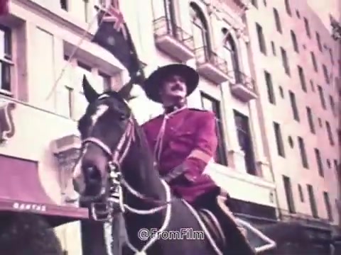 The still from the 16mm film 'Canada Dry Ginger Ale - The Unexpected Soft Drink 1970's' features a mounted figure in a distinct red uniform, likely representing a member of the Royal Canadian Mounted Police. The background showcases a blend of historic architecture and flags, creating a nostalgic atmosphere reflective of Canadian culture. This scene captures a moment of celebration or community engagement, emphasizing the connection between the drink and a sense of national pride.