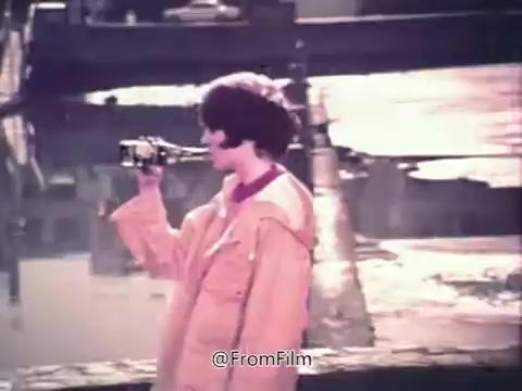 The still from the 16mm print of 'Canada Dry Ginger Ale - The Unexpected Soft Drink' features a woman in a light orange rain jacket enjoying a bottle of Canada Dry ginger ale. The setting appears to be outdoors, possibly near water or a dock, and the visual style evokes a nostalgic 1970s aesthetic. The focus is on the enjoyment of the drink, suggesting a refreshing and surprising experience.