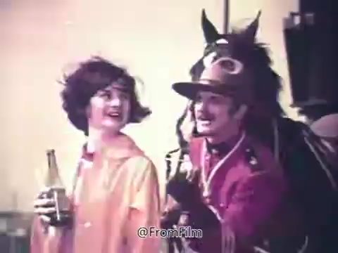 The still from the 1970s Canada Dry Ginger Ale commercial features a woman in a bright, stylish outfit holding a bottle of ginger ale, smiling cheerfully. Beside her is a man dressed in a classic Mountie outfit, exuding charm and playfulness. A horse can be seen in the background, adding to the whimsical and light-hearted atmosphere of the scene. The overall vibe captures the era's fun and unexpected take on soft drinks.