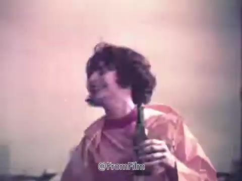 The still from 'Canada Dry Ginger Ale - The Unexpected Soft Drink' features a person with curly hair, joyfully holding a bottle, wearing a colorful raincoat. The background appears hazy, perhaps suggesting a whimsical or surreal setting, typical of 1970s advertising. The overall tone conveys a sense of fun and spontaneity associated with the brand.
