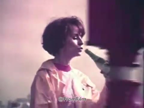 The still from the 1970s Canada Dry Ginger Ale commercial features a woman in a light-colored rain jacket holding a bottle of ginger ale. She appears to be enjoying the drink, with a contemplative expression, suggesting a refreshing and unexpected experience. The background is softly blurred, giving a dreamy, nostalgic feel typical of that era’s advertising aesthetic. The vibrant colors and casual setting evoke a sense of comfort and enjoyment associated with the product.