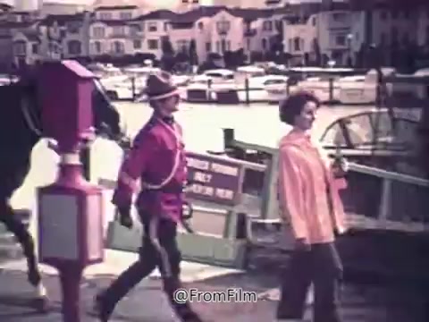The still from 'Canada Dry Ginger Ale - The Unexpected Soft Drink 1970's' features a Mountie in traditional uniform walking alongside a woman near a marina. The background shows boats and a row of houses, capturing a scenic and leisurely atmosphere typical of the era. The image emphasizes a sense of community and Canadian identity, aligning with the branding of Canada Dry Ginger Ale.