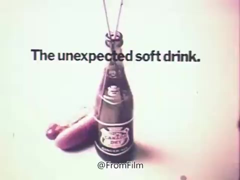 The still features a Canada Dry Ginger Ale bottle prominently displayed alongside a couple of hot dogs. The text 'The unexpected soft drink' overlays the image, emphasizing the product's unique position in the soft drink market during the 1970s. The color palette reflects the era's aesthetic, with a vintage feel characteristic of 16mm prints. This advertisement aims to convey the refreshing nature of Canada Dry in a playful, unexpected context.