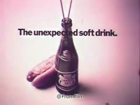 The still features a bottle of Canada Dry Ginger Ale, depicted as an unexpected choice for a soft drink. The bottle is centrally placed, with two straws inserted, suggesting a whimsical approach to drinking. The phrase “The unexpected soft drink” is prominently displayed above the bottle, emphasizing the brand's unique positioning in the soda market during the 1970s. The background is likely simple to keep focus on the product.