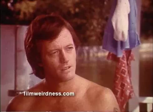 The still from 'Outlaw Blues' features a shirtless man, portrayed by Peter Fonda, sitting outdoors with a contemplative expression. The background suggests a relaxed, summer setting, likely near water, with clothing hanging nearby. The film explores themes of rebellion and music, characteristic of Fonda's iconic roles during that era.