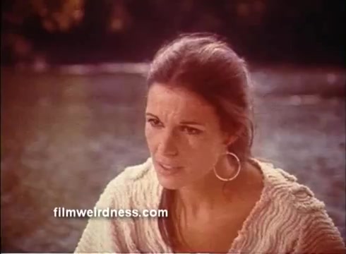 The still from the 'Outlaw Blues' trailer features a woman with long, flowing hair and large hoop earrings, expressing concern or intensity. She is dressed in a light, textured outfit, with a natural, outdoor backdrop indicating a moment of emotional significance. The overall tone suggests a blend of drama and personal stakes related to the film's plot.