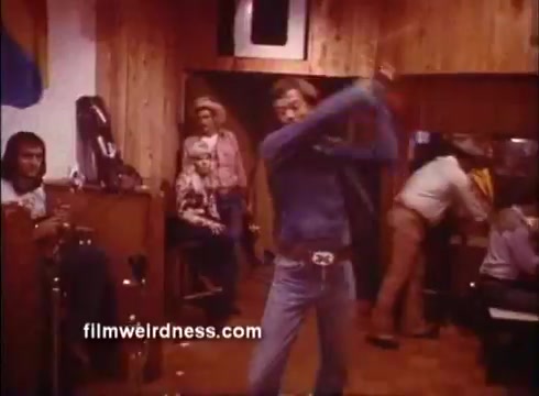 The still from the 'Outlaw Blues Trailer' features a dynamic scene set in a rustic bar environment. A man, presumably Peter Fonda's character, is depicted in the foreground, swinging a baseball bat with intensity. The background includes a few onlookers, some appearing surprised, while two individuals are engaged in conversation. The wooden decor and casual attire of the characters hint at a laid-back, yet tense atmosphere, typical of a classic Western-themed film.