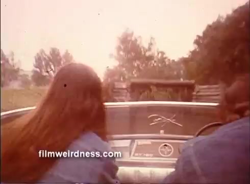 The still features a view from the inside of a convertible car, with a focus on the dashboard and the exterior ahead. A couple of individuals, one with long hair, is captured as they appear to be driving in a rural area, possibly engaged in a moment of adventure. The scene has a warm, vintage tone typical of 1970s films, enhancing the nostalgic atmosphere of 'Outlaw Blues.'