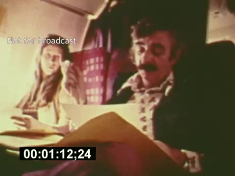 The still from 'Sixty Second Spot' depicts a moment on an airplane during the mid-1970s, featuring a man and a woman engaged in discussion. The man, dressed in a patterned shirt, appears to be reviewing a script or materials for a 7-Up commercial, while the woman listens attentively. The vintage aesthetic highlights the era's filmmaking style and offers insight into the behind-the-scenes processes of creating a TV commercial. The timestamp indicates it’s an excerpt focused on script development and casting, setting the stage for the eventual filming of the ad.