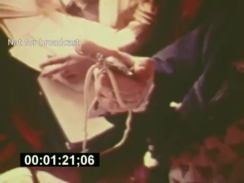 The still from 'Sixty Second Spot' depicts a behind-the-scenes moment in the creation of a 7-Up commercial from the mid-1970s. The focus is on a hand holding a small puppet or prop, possibly related to the commercial's theme. Script notes and other materials are visible in the background, highlighting the collaborative process of casting, rehearsing, and filming. The timestamp indicates it occurs early in the film, emphasizing the development stage of the advertisement.