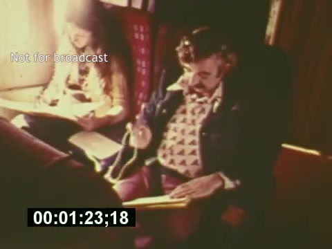 The still from 'Sixty Second Spot' shows a creative setting where a man and a woman are working on the production of a 7-Up commercial. The man is seated with a notepad, focused on his task, while the woman appears to be engaged in similar work nearby. The relaxed, vintage atmosphere reflects the mid-1970s era of advertising, highlighting the behind-the-scenes process from script development to casting and filming. A timestamp indicates a moment in the film, likely emphasizing the creative process involved in making the commercial.