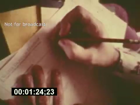 The still from 'Sixty Second Spot' captures a close-up of hands writing on a piece of paper, likely part of the script development process for a 7-Up commercial. It highlights the behind-the-scenes effort involved in creating a TV commercial during the mid-1970s. The image includes a timestamp and a 'Not for broadcast' label, emphasizing its documentary nature and context within the film.