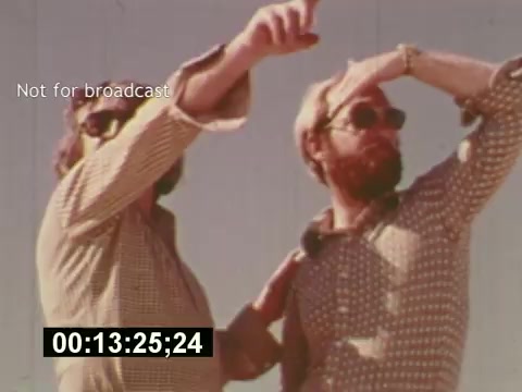 The still from 'Sixty Second Spot' depicts two men outdoors, likely during the production of a 7-Up commercial in the mid-1970s. They are engaged in discussion, gesturing and pointing as they collaborate on the project. Both wear sunglasses, suggesting a sunny setting, and their attire reflects the casual fashion of the era. This moment captures the behind-the-scenes process of commercial creation, showcasing the creative energy involved in bringing a script to life.