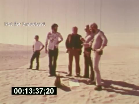 The still features a group of five men standing in a sandy, desolate location, likely a desert, as they engage in discussion about the creation of a 7-Up commercial. They are dressed in casual 1970s attire, and the scene conveys a collaborative atmosphere of the filmmaking process. The timestamp indicates it's part of a video, marking a moment in the mid-1970s film, 'Sixty Second Spot,' which explores the stages from script development to casting and filming. The presence of the 'Not for broadcast' label suggests it is intended for internal or archival purposes, not for public viewing.