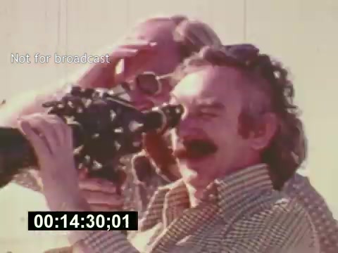 The still depicts a scene from 'Sixty Second Spot,' showcasing the creation of a 7-Up commercial in the mid-1970s. It features a man with a mustache operating a film camera while another individual appears to shield their eyes from the sun. This moment captures the behind-the-scenes effort of filmmaking, emphasizing the creative process involved in commercial production. The timestamp indicates a specific moment in the film, adding context to the ongoing sequence of filming.