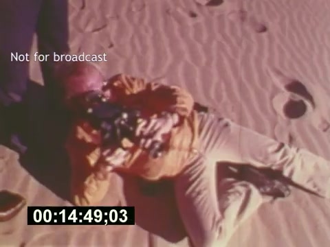 The still from 'Sixty Second Spot' captures a moment during the production of a 7-Up commercial in the mid-1970s. It features a crew member lying on sandy terrain, presumably adjusting equipment or taking a break during filming. The scene emphasizes the behind-the-scenes efforts involved in commercial creation, showcasing the informal and dynamic aspects of film production. The watermark 'Not for broadcast' indicates that this is a production still rather than a publicly released image. The timestamp suggests it's part of a timeline in the film, highlighting the process of bringing the advertisement to life.