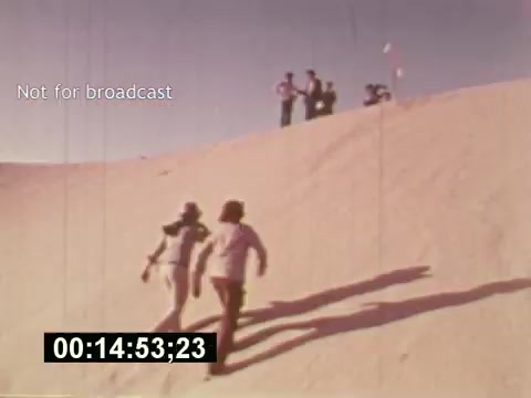 The still from 'Sixty Second Spot' captures a moment of the filmmaking process for a 7-Up commercial from the mid-1970s. In the scene, two people are running up a sandy hill, likely portraying the energetic vibe of the brand. In the background, a small crew can be seen observing the action, emphasizing the collaborative nature of commercial production. The timestamp and 'Not for broadcast' label indicate it's an unedited part of the film, showcasing behind-the-scenes creativity.
