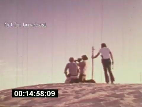 The still from 'Sixty Second Spot' captures a behind-the-scenes moment in the creation of a 7-Up commercial from the mid-1970s. It shows a small crew on location, with one person adjusting a camera while others observe the setup against a sandy backdrop. The scene illustrates the collaborative effort involved in filmmaking, emphasizing the nuances of commercial production during that era. The timecode and 'Not for broadcast' label indicate its status as a production still.