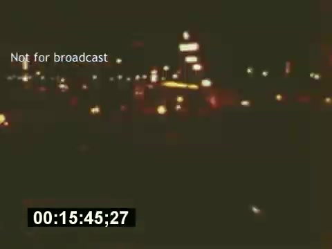 The 16mm still from 'Sixty Second Spot' captures a nighttime scene, likely during the filming of a 7-Up commercial. The image portrays a bustling urban environment, illuminated by streetlights and neon signs, setting a vibrant backdrop for the ad's creation process. The timestamp in the lower corner indicates a specific moment in the film, emphasizing its candid exploration of the behind-the-scenes efforts involved in commercial production.