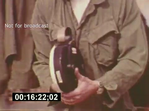 The still captures a moment from the film 'Sixty Second Spot,' showcasing a behind-the-scenes look at the creation of a 7-Up commercial. In this frame, a person dressed in an olive-green shirt is holding a piece of filming equipment, likely a camera or sound device. The visual emphasizes the craftsmanship and effort involved in producing a television advertisement, indicative of the mid-1970s film production style. A timestamp indicates the scene's position within the film, enhancing its documentary feel.