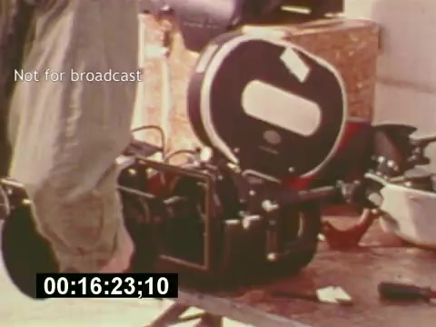 The still from 'Sixty Second Spot' features a 16mm film camera set up for the creation of a 7-Up commercial. The shot captures a moment in the filmmaking process, showcasing the equipment used in the mid-1970s, likely during a scene of preparation or filming. The timestamp indicates the film's progression, emphasizing the behind-the-scenes aspect of commercial production from scripting to casting and on-set activities.