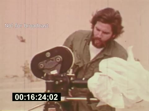 The 16mm still from 'Sixty Second Spot' depicts a man with a beard operating a film camera, capturing the process of creating a 7-Up commercial. He's dressed in a casual, olive-green shirt, and the backdrop appears to be a sandy or outdoor location. This moment likely highlights the technical aspects of filmmaking in the mid-1970s, focusing on the behind-the-scenes work leading up to the final commercial product. The timestamp suggests it's from an ongoing scene in the film.