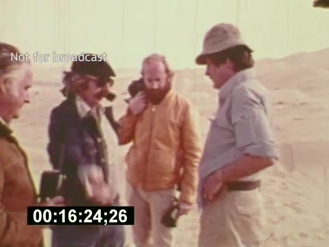 The still from 'Sixty Second Spot' captures a moment during the production of a 7-Up commercial in the mid-1970s. It features a group of four men, likely including the director and crew, engaged in discussion on location, potentially in a desert setting. The visible elements suggest a collaborative atmosphere as they navigate the creative process of filmmaking, with a time code overlay indicating where this moment occurs in the film. The title 'Not for broadcast' implies this is a behind-the-scenes glimpse into the making of the commercial.