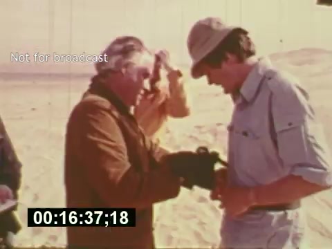 The still from 'Sixty Second Spot' captures a behind-the-scenes moment in the creation of a 7-Up commercial during the mid-1970s. It features a group of individuals on a sandy set, indicating a remote filming location. Two men, one wearing a hat and another in a brown jacket, appear to be working closely together, possibly discussing camera equipment or directions. The image highlights the collaborative nature of commercial filmmaking, detailing the casting and production process as the project unfolds. The on-screen timer implies a focus on specific timing within the film.