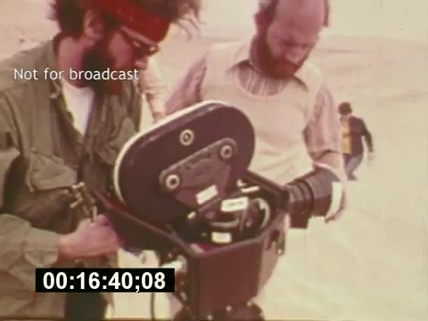 The still captures a behind-the-scenes moment from the film 'Sixty Second Spot,' showcasing the production of a 7-Up commercial from the mid-1970s. Two men are seen examining a 16mm film camera, indicating the on-location shooting process. The setting appears to be an outdoor environment, possibly a desert or open field, hinting at the film's creative atmosphere. Text overlay states 'Not for broadcast,' suggesting this is exclusive production footage. The timestamp at the bottom indicates a specific moment in the film.