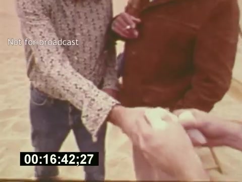 The still from 'Sixty Second Spot' captures a moment during the creation of a 7-Up commercial from the mid-1970s. In the frame, two individuals are engaged in a discussion, likely about the production process. The scene highlights the collaborative effort involved in filmmaking, showcasing hands exchanging something—possibly a script or prop—against a sandy backdrop. The timecode suggests this is a specific moment within the film, emphasizing the behind-the-scenes nature of commercial creation.