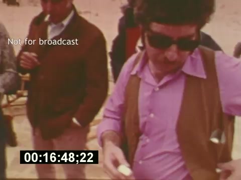 The still from 'Sixty Second Spot' captures a behind-the-scenes moment from the production of a 7-Up commercial in the mid-1970s. It shows a man wearing sunglasses and a pink shirt with a brown vest, seemingly checking something in his hand. In the background, other crew members are engaged in conversation, hinting at the collaborative nature of film production. A time code in the corner indicates the moment in the footage, and the 'Not for broadcast' label suggests this is a private or developmental shot rather than a final product. This image encapsulates the creative process and teamwork involved in making a TV commercial.