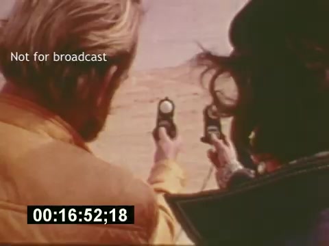 The still from *'Sixty Second Spot'* depicts two characters aiming guns in a desert setting, likely part of a scene intended for a 7-Up commercial. The actors are positioned closely, with the viewer seeing their hands and the guns prominently. The film, from the mid-1970s, showcases the creative process of making a TV commercial, including script development, casting, and filming, culminating in the final advertisement. The timestamp suggests this moment is part of the behind-the-scenes footage leading to the completed commercial.