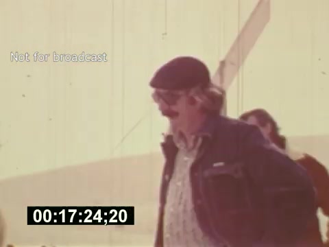 The still from 'Sixty Second Spot' captures a moment during the production of a 7-Up commercial in the mid-1970s. It features a man with a mustache and a beret walking casually, indicating a behind-the-scenes atmosphere. The faded quality of the film suggests an authentic period look, emphasizing the film's exploration of the commercial creation process from script to casting to filming. The timestamp indicates it’s part of the editing timeline, hinting at the emphasis on production details and the challenges faced during the shoot.