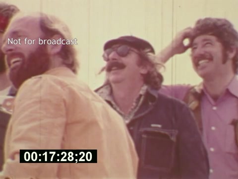 The still from 'Sixty Second Spot' captures a moment from the mid-1970s during the making of a 7-Up commercial. Three men, dressed in casual clothing, are smiling and laughing, reflecting a lighthearted atmosphere on set. One man wears sunglasses and a hat, while another has a noticeable mustache. The background appears bright and slightly blurred, emphasizing their expressions. The time code on the bottom indicates that this is a specific moment in the film, and the 'Not for broadcast' label suggests it's a behind-the-scenes glimpse rather than the finished commercial.