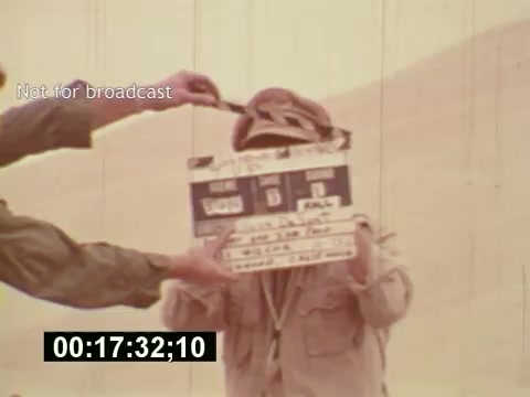 The still captures a moment from the film 'Sixty Second Spot,' highlighting the behind-the-scenes process of creating a 7-Up commercial from the mid-1970s. In the image, a crew member holds a clapperboard to mark the start of a take, while another person, dressed in casual attire, stands ready for filming. The background suggests an outdoor setting, contributing to the documentary style. The timestamp indicates the specific moment in the film when this shot occurs.