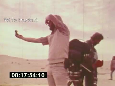 The still from 'Sixty Second Spot' captures a scene in a desert setting during the production of a 7-Up commercial. The image features a crew member holding a miniature model or prop, possibly evaluating its placement or scene composition. Behind him, another crew member is seen carrying equipment, indicating the hands-on nature of filmmaking in the mid-1970s. The overall mood reflects the collaborative process involved in commercial production, showcasing both the creative and logistical aspects of bringing an advertisement to life. The timestamp and 'Not for broadcast' label highlight its purpose as an archival piece rather than a public airing.