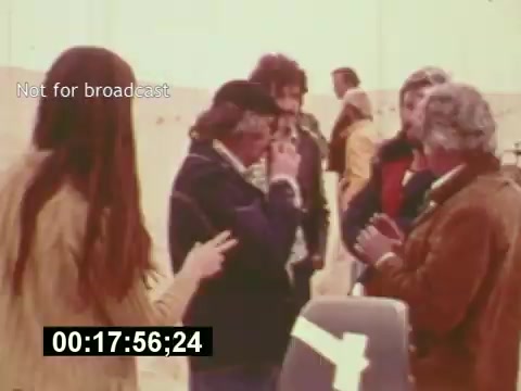 The still from 'Sixty Second Spot' captures a behind-the-scenes moment in the creation of a 7-Up commercial from the mid-1970s. Featured are a group of filmmakers and actors engaged in discussion, likely brainstorming or preparing for a scene. The setting appears casual, with the participants dressed in vintage fashion typical of the era. A camera setup is visible, indicating the filming process is underway. The timecode and 'Not for broadcast' label suggest it is a raw or preliminary clip from the documentary.