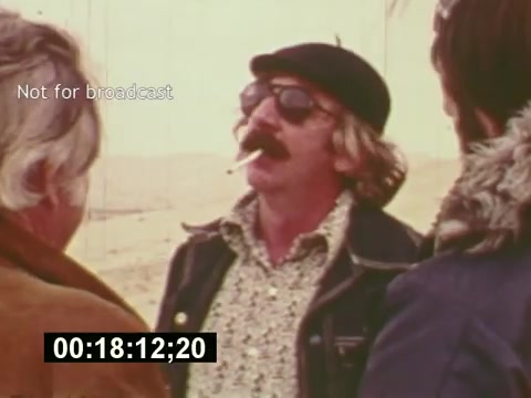 The still from 'Sixty Second Spot' depicts a moment during the creation of a 7-Up commercial in the mid-1970s. It features a character wearing sunglasses and a hat, exuding a laid-back vibe, with a cigarette in hand. Surrounding him are two individuals engaged in conversation, likely discussing aspects of the commercial's production. The sandy backdrop suggests an outdoor setting, possibly reflecting the filming location. This behind-the-scenes glimpse captures the casual atmosphere of the era and the creative process of advertising.