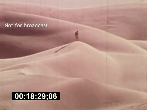 The still from 'Sixty Second Spot' features a lone figure traversing sand dunes, highlighting the expansive and barren landscape characteristic of desolate desert scenes. The visual focuses on the interplay of light and shadow on the dunes, creating a warm, soft aesthetic typical of 1970s cinematography. The text 'Not for broadcast' indicates that this image is part of behind-the-scenes footage rather than intended for public airing, and the timecode suggests it is from the film’s playback format. This moment captures the contemplative tone of the film, which chronicles the process behind producing a 7-Up commercial.
