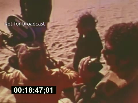 The still from 'Sixty Second Spot' captures a behind-the-scenes moment in the making of a 7-Up commercial from the mid-1970s. It features crew members, including a cinematographer with a camera, and others involved in the production discussing or preparing for a scene. The visual hints at the collaborative atmosphere of filming, set against a sandy backdrop that suggests an outdoor setting. The accompanying timestamp indicates it’s a precise moment within the film's narrative, which details the entire creative process of commercial production.