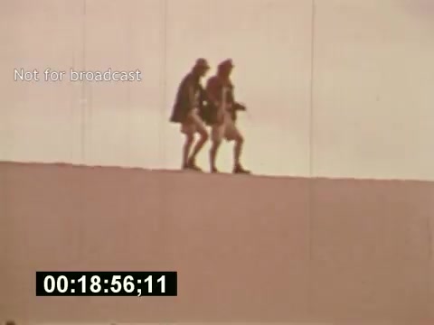 The still from 'Sixty Second Spot' depicts two individuals walking along a sandy surface, likely on location for a 7-Up commercial shoot from the mid-1970s. The scene captures the behind-the-scenes process, showcasing the creative journey from script to filming. The tone is nostalgic, reflecting the filmmaking style of that era. The overlay 'Not for broadcast' suggests its exclusive nature, emphasizing its purpose as a documentary insight rather than a finished advertisement. The timecode indicates it’s part of the film’s runtime.