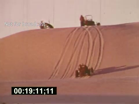 The still from 'Sixty Second Spot' captures a scene depicting the production of a 7-Up commercial in the mid-1970s. In this moment, vehicles are seen maneuvering on a sandy landscape, leaving distinct tire tracks. This part of the film showcases the behind-the-scenes aspects of commercial creation, focusing on elements like casting and filming techniques. The timestamp at the bottom indicates its location within the overall narrative of the project.