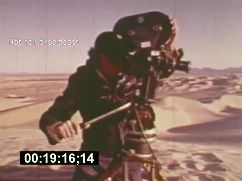 The still from 'Sixty Second Spot' captures a behind-the-scenes moment of the 7-Up commercial production in the mid-1970s. It shows a crew member operating a 16mm camera, set against a spacious desert landscape. The image highlights the technical aspects of filmmaking during that era, with a focus on the meticulous process of casting and filming. A timestamp is visible at the bottom, emphasizing the documentary style of the film, which chronicles the creation of the commercial from script to screen.