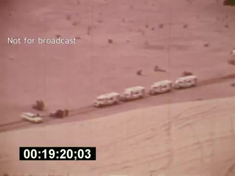 The still from 'Sixty Second Spot' depicts a wide aerial view of a desert landscape where several production vehicles are lined up along a dirt road. The scene captures the essence of film production in the mid-1970s, showcasing behind-the-scenes elements involved in creating a 7-Up commercial. The color palette reflects the era's film style, and the overlay indicates that the footage is not intended for broadcast.